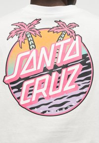 Valkoisen paidan takapuoli, jossa on värikäs pyöreä logo, jossa kaksi vaaleanpunaista palmuparvea, auringonlaskun väreissä ja teksti "Santa Cruz Skateboards".