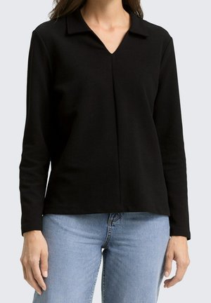 Femme portant un haut noir à manches longues avec col en V et un jean bleu clair, debout devant un fond clair uni.
