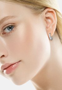 Orecchino a cerchio in argento con una finitura liscia e lucida, caratterizzato da una curva sottile e un leggero dettaglio di design. Indossato sull'orecchio di un modello.