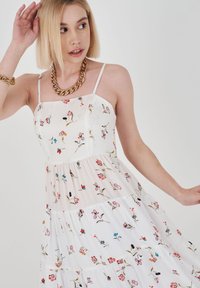 Blumenbedrucktes, gestuftes Kleid aus weißem Stoff mit dünnen Trägern und einem taillierten Oberteil. Akzente setzen bunte Blumendesigns.
