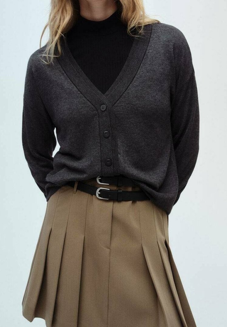 Femme portant un cardigan gris foncé boutonné sur un haut noir, associé à une jupe plissée beige et une ceinture noire à double boucle.