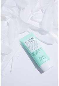 Mintgrön tub med Benton UV Defense Solkräm, med vit text och glansig yta, placerad på mjuka, vita fjädrar.