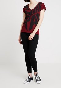 Burgundy v-ringad t-shirt med svart geometrisk tryck, ihopparad med svarta slim-fit jeans och svarta sneakers. Enkel design, bomullsmaterial.