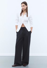 Chemise blanche à col boutonné avec un design cropped et des manches longues, associée à un pantalon noir à rayures fines taille haute et large. Chaussures marron.