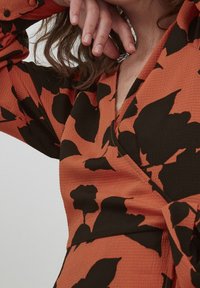 Robe portefeuille en tissu orange rouille avec une surface texturée, présentant de grands motifs floraux noirs et des manches longues. Détail à encolure en V visible.