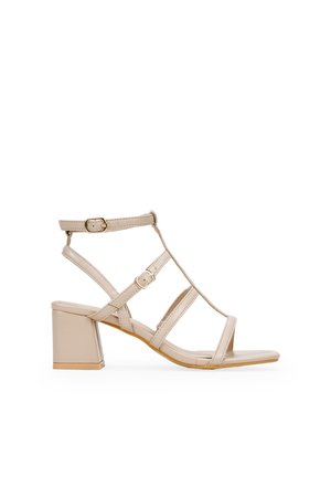 Sandalia de tacón beige con tiras y tacón cuadrado. Presenta múltiples tiras ajustables y una superficie lisa y brillante. Diseño de punta abierta y plana.