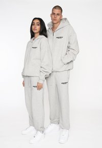 Conjunto de sudadera y pantalones de chándal en gris claro, con un logo en el pecho y en los pantalones. La tela es suave y tiene un corte relajado. Zapatillas blancas completan el look.