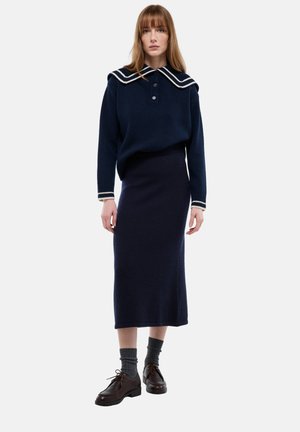 Pull en maille bleu marine avec un col marin, orné de bandes blanches contrastantes. Associé à une jupe ajustée bleu marine, des chaussettes grises et des chaussures marron.