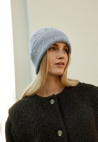 Grijze gebreide muts met een geribbelde textuur, gedragen met een zwarte, zachte jas met ronde knopen, tegen een neutrale achtergrond.
