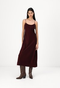 Rochie maxi din satin burgund cu bretele subțiri și o croială lejeră, purtată cu cizme cowboy cu toc maro. Textură fină și design minimalist.