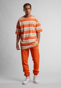 Übergroßes T-Shirt mit orange-beigen Streifen und kurzen Ärmeln, kombiniert mit orangen Sweatpants mit einem weißen Logo und weißen Sneakers.