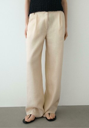 Pantalon beige à jambes larges porté avec des tongs noires et un haut noir en tricot sans manches, montré de la taille aux pieds.