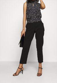 Ärmlös svart topp med vit prickmönster och nyckelhålsringning, matchad med smala svarta byxor och svarta klacksandaler.