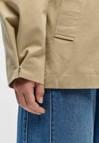 Main droite détendue sur le côté, portant une manche de veste beige et un jean en denim bleu, montrant la texture du tissu et les détails des coutures.