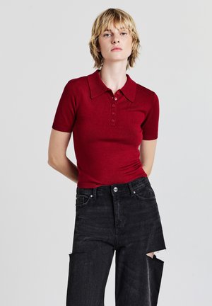 Frau mit kurzen blonden Haaren, die ein enganliegendes rotes Polohemd mit kurzen Ärmeln und schwarze weite Jeans mit großen Knieausschnitten trägt.