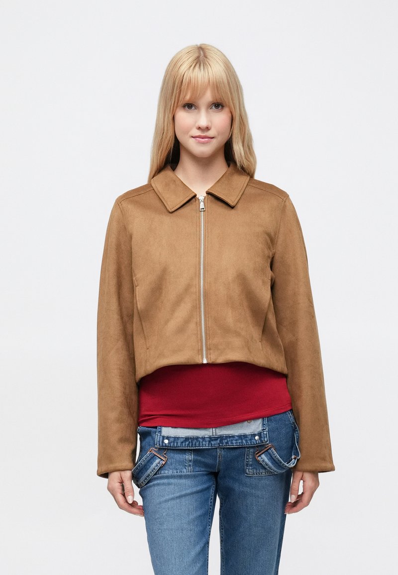 Brun ruskind cropped jakke med krave, med en frontlynlås og lange ærmer, båret over en rød top og denimjeans.