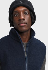 Giacca in pile blu navy con colletto alto e maniche a maglia grigia in contrasto. Presenta una zip frontale e un cappellino di lana a coste nero.