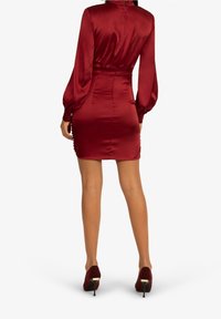 Czerwono-satynowa sukienka z długimi bufkami, wcięciem w talii i krótkim hemem. Dobrana z bordowymi szpilkami. Widok z tyłu.