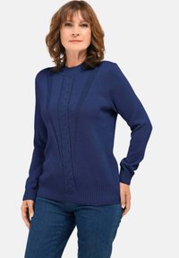 Vrouw in een marineblauwe long sleeve trui met textuur verticale en geometrische breipatronen, gecombineerd met een blauwe jeans, staande tegen een effen achtergrond.