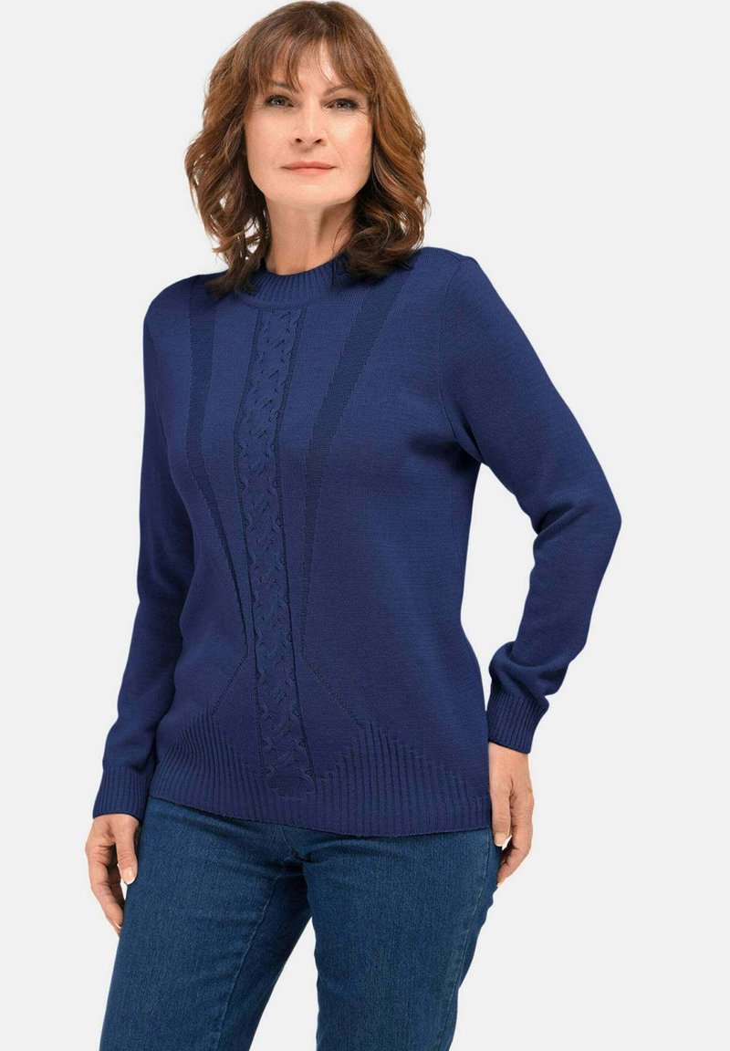 Vrouw in een marineblauwe long sleeve trui met textuur verticale en geometrische breipatronen, gecombineerd met een blauwe jeans, staande tegen een effen achtergrond.