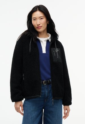 Ženska z dolgimi temnimi lasmi nosi črno flis jakno čez modro in belo srajco z jeans hlačami na belem ozadju.