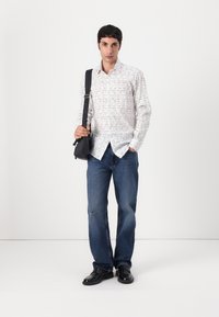 Hvit skjorte med svart tekstmønster, kombinert med blå jeans og svarte sko. Modellen holder en svart crossover-veske.