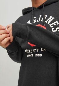Schwarzer Kapuzenpullover mit langen Ärmeln, gerippten Bündchen und weißem Text. Verfügt über ein rotes Logo auf der Brust und eine verstellbare Kapuze mit Kordelzug.