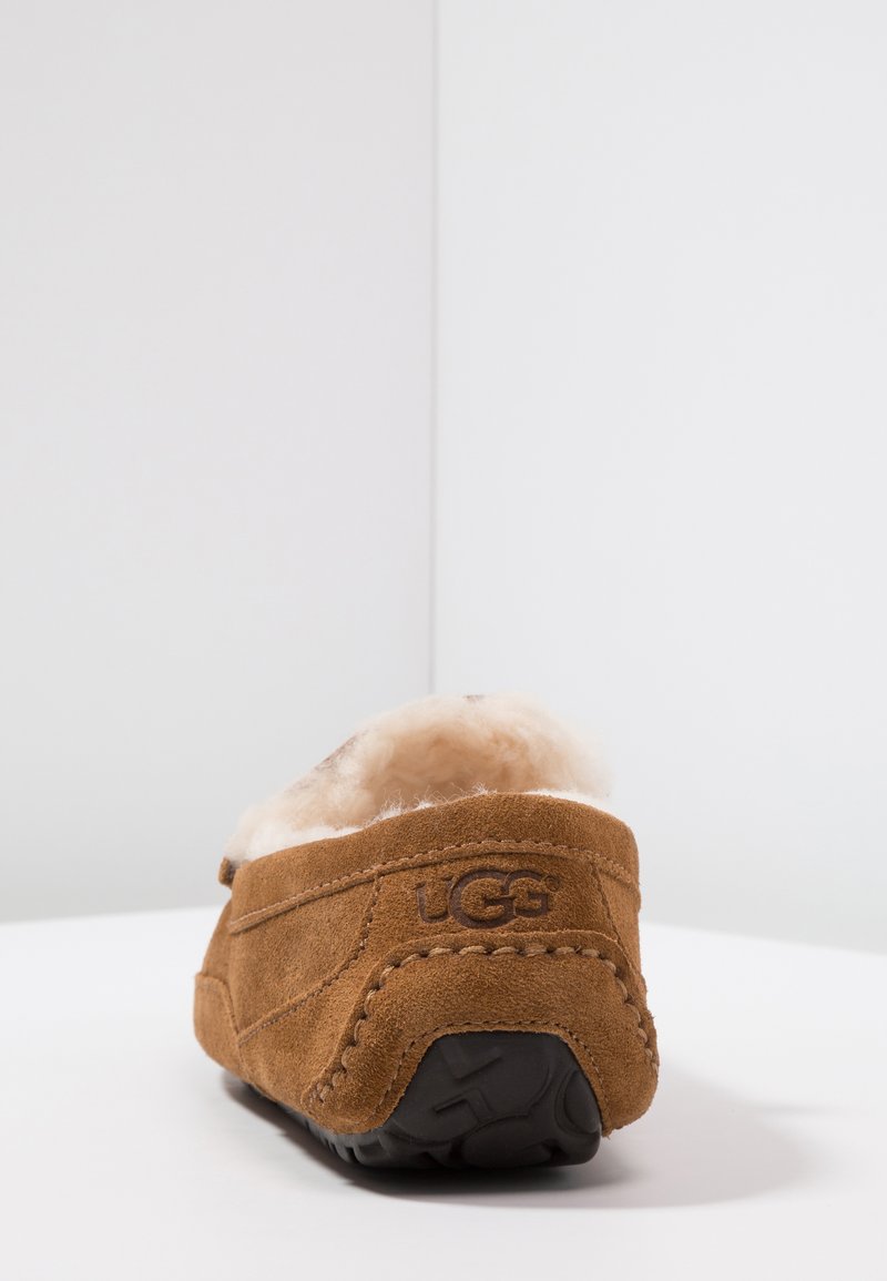 Brown suede slipper with plush interior lining, rubber sole, and embroidered logo. Features a smooth texture and rounded back design.