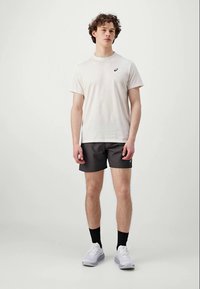 Junger Mann, der ein weißes kurzärmliges Hemd, schwarze Shorts, schwarze Socken und weiße Sportschuhe trägt, vor einem einfarbigen Hintergrund steht.