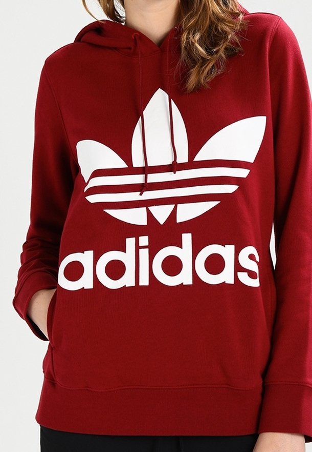 Červená mikina s kapucí vyrobená z bavlněné směsi, s velkým bílým logem Adidas a třemi vodorovnými pruhy na přední straně.