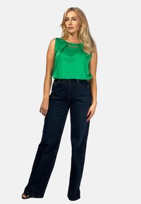 Zielona bezrękawnikowa bluzka z łańcuszkowym akcentem przy dekolcie, połączona z ciemnoniebieskimi spodniami dzwonami. Gładki materiał i luźny krój.