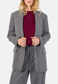 Blazer grigio oversize con vestibilità sartoriale, revers a punta e tasche laterali, indossato sopra un top borgogna, abbinato a pantaloni grigi coordinati.