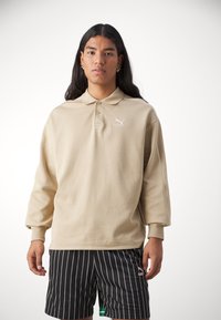 Beige Poloshirt mit langen Ärmeln aus einem strukturierten Stoff, ausgestattet mit einer Zwei-Knopf-Leiste und einem kleinen weißen Puma-Logo auf der linken Brust.