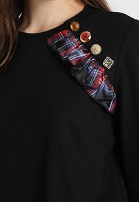 Svart sweatshirt med en vågig rand i röd, blå och svart, prydd med tre dekorativa knappar på axeln.