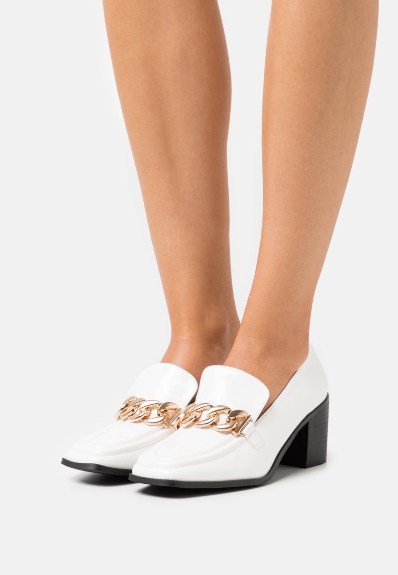 Mocassins en cuir blanc à talons blocs avec un détail de chaîne dorée, portés sur des jambes nues contre un fond uni.