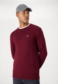 Pull en maille bordeaux avec un col rond, des poignets et un ourlet côtelés, arborant un petit logo sur la poitrine. Porté avec une casquette grise.