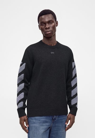 Jeune homme portant un pull noir avec des rayures diagonales grises sur les manches et un petit texte "OFF" sur la poitrine, associé à un jean bleu, sur fond blanc.