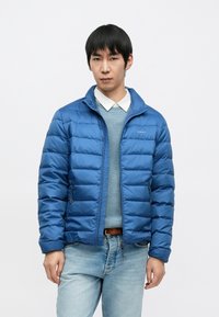 Geacă puf albastră cu fermoar frontal, cu guler tip în picioare și model matlasat orizontal. Purtați peste un pulover albastru deschis și blugi.