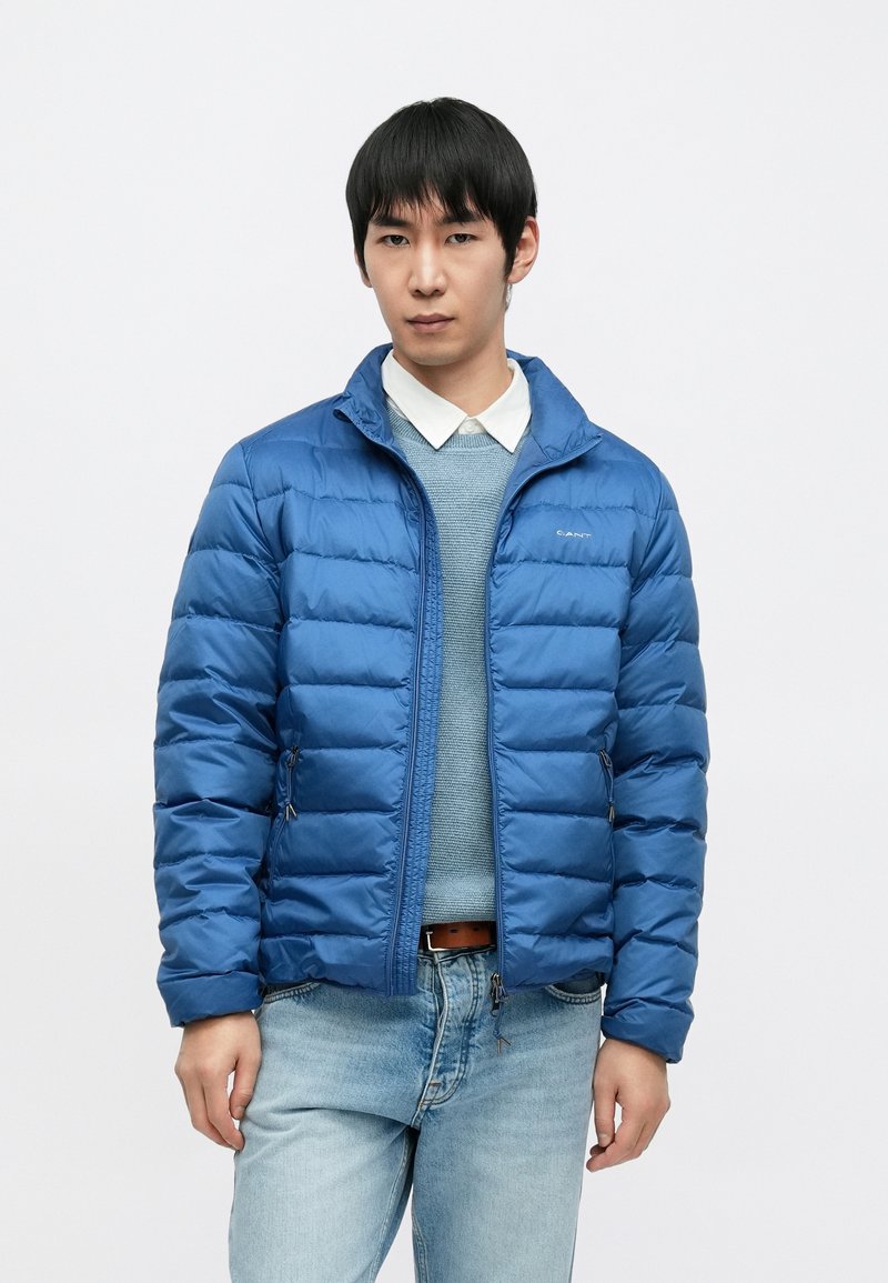 Geacă puf albastră cu fermoar frontal, cu guler tip în picioare și model matlasat orizontal. Purtați peste un pulover albastru deschis și blugi.