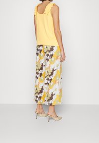 Geel mouwloos topje met gerimpelde halslijn, gecombineerd met wijde broek in een bloemmotief in bruin, geel en wit. Beige hakken.