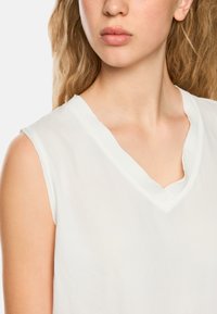 White sleeveless blouse with a soft texture, featuring a V-neckline and clean edges, showing minimal fabric draping.