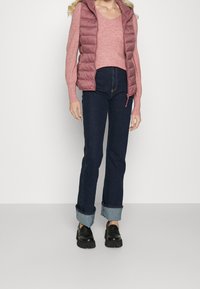 Kobieta w różowym swetrze, lawendowej pikowanej kamizelce, ciemnych dżinsach z mankietami i czarnych, masywnych loafersach stoi na tle jednorodnym.