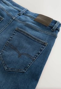 Tummansiniset farkut, joissa on ruskea nahkapaikka vyötäröllä. Farkuissa on yksi takatasku, jossa on kaarevaa ompeluompelua.