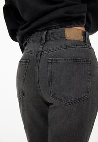 Black denim jeans with a slightly faded texture, featuring a tan leather patch at the waistband and two back pockets.