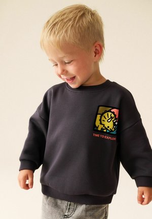 Ung blond dreng iført en mørk sweatshirt med en farverig dinosaur-grafik og teksten "Time to Explore", kigger ned og smiler.