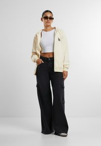 Cremefarbener Zip-Hoodie, enganliegendes weißes Crop-Top und schwarze weit geschnittene Cargo-Hosen mit Seitentaschen. Das Modell trägt eine dunkle Sonnenbrille.