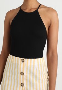 Černý žebrovaný top na krk s tenkými popruhy, nošený s žlutou a bílou pruhovanou sukní, která má velké knoflíky vpředu.