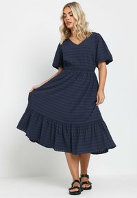 Sinivihreä ruudullinen mekko V-päällä, lyhyet puffihihat, elastinen vyötärö ja liehuva, röyhelöreuna. Yhdistetty mustiin sandaaleihin.