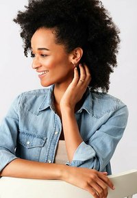 Camicia in denim di un blu chiaro con bottoni metallici, indossata sopra un top beige. Modella con capelli ricci neri e piccoli orecchini a perno, sorridente.