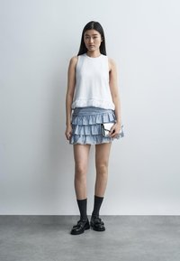 Femme portant un haut sans manches blanc, une mini-jupe bleue à volants, des chaussettes noires, des mocassins, tenant une petite pochette métallique, debout contre un mur uni.